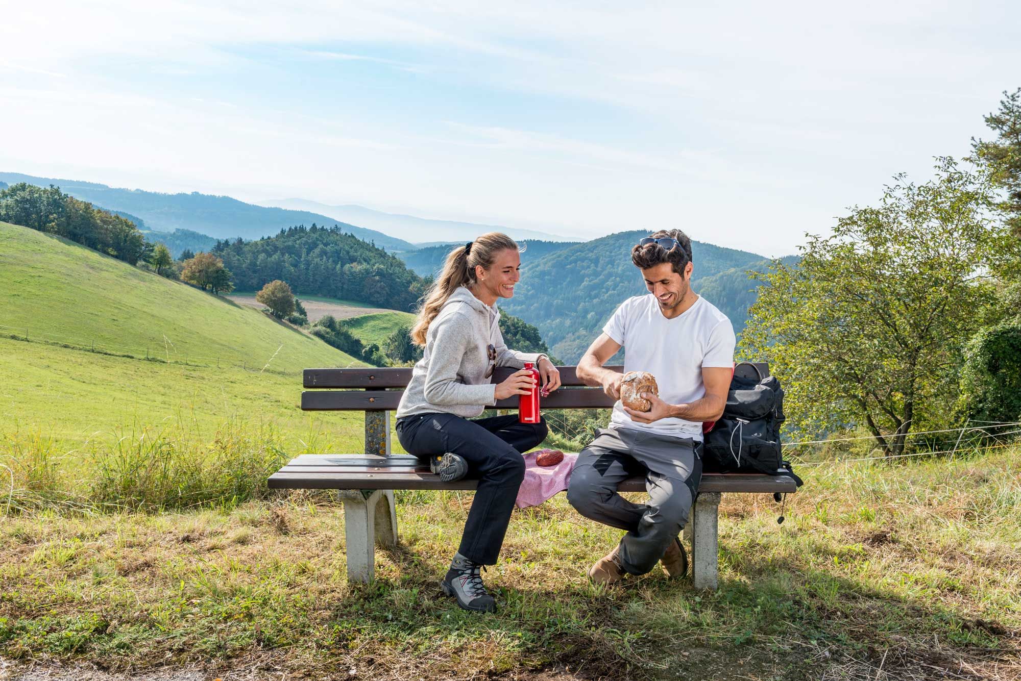 Zwei Personen sitzen auf einer Bank in einer hügeligen Landschaft und machen eine Pause beim Wandern.