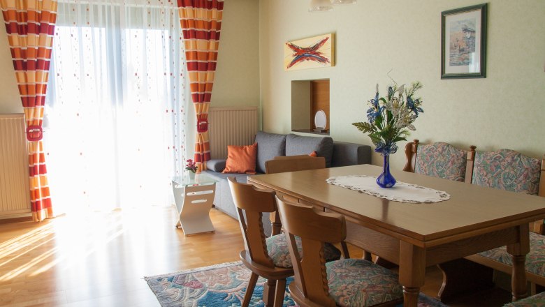 Wohnzimmer, © Fam. Sulzbacher Ein gemütliches Wohnzimmer mit Holztisch, Stühlen, grauem Sofa und bunten Vorhängen. Ein Blumenstrauß steht auf dem Tisch.