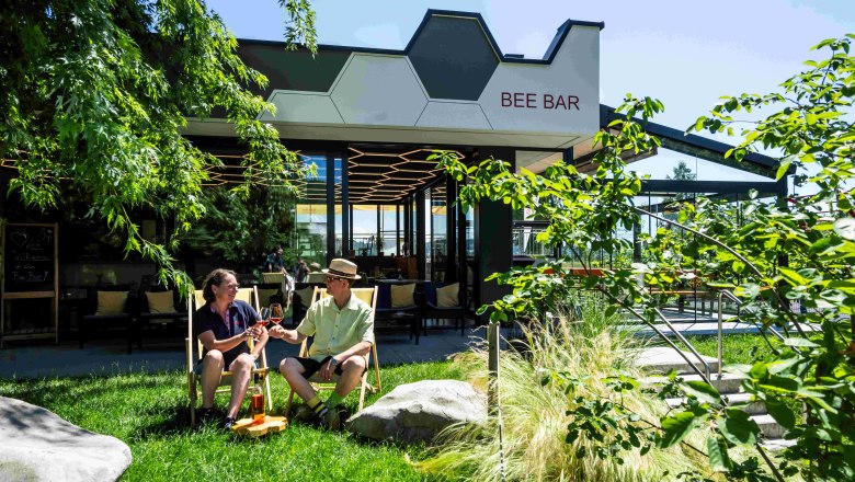 Zwei Personen sitzen im Garten vor der Bee Bar und stoßen mit Getränken an.