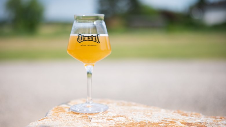 Ein Glas mit hellem Bier und Braustall-Logo steht auf einem Stein im Freien.