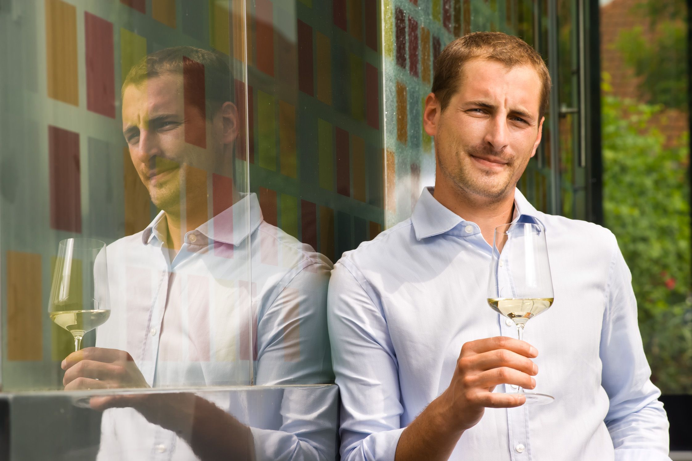 Ein Mann in einem hellblauen Hemd hält ein Glas Weißwein und lehnt an einer bunten Glaswand.