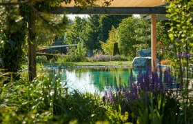 Ein idyllischer Garten mit Teich, umgeben von üppiger Vegetation und blühenden Pflanzen, unter einem schattenspendenden Pavillon.