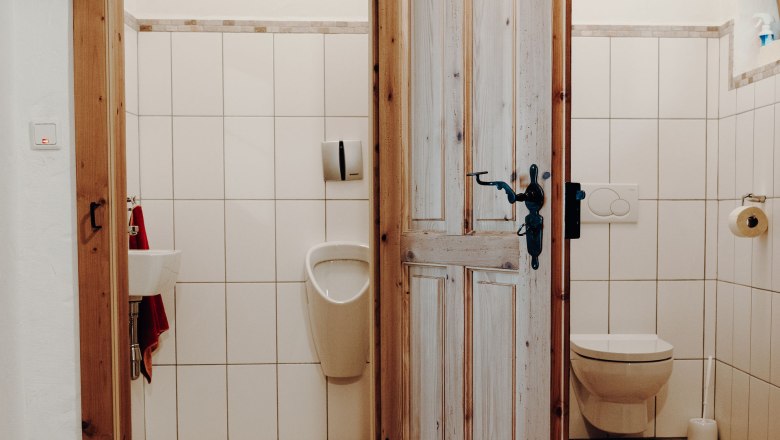 Zwei Toilettenr&auml;ume mit Holzt&uuml;r, links ein Urinal und Waschbecken, rechts eine Toilette.