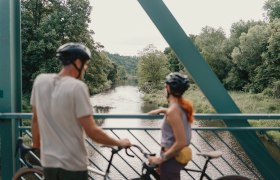 Zwei Radfahrer mit Helmen stehen auf einer Brücke und blicken auf einen Fluss, umgeben von üppiger Vegetation.