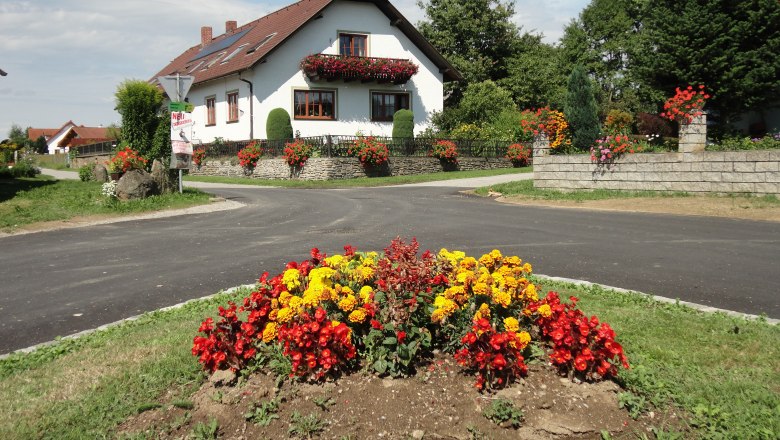Ein weißes Haus mit roten Dachziegeln und Blumenschmuck, umgeben von bunten Blumenbeeten und grüner Landschaft.
