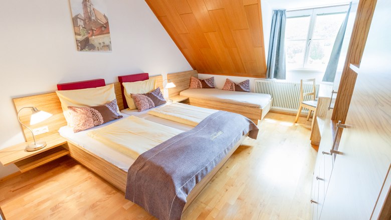 Gem&uuml;tliches G&auml;stezimmer mit Doppelbett, Einzelbett, Holzverkleidung und gro&szlig;em Fenster.