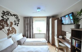 Ein modernes Schlafzimmer mit Doppelbett, Wanddekoration, Schreibtisch, Fernseher und Pflanzen.