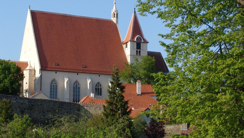 Pfarrkirche St. Stephan mit rotem Dach und Turm, umgeben von Bäumen und einer Steinmauer.
