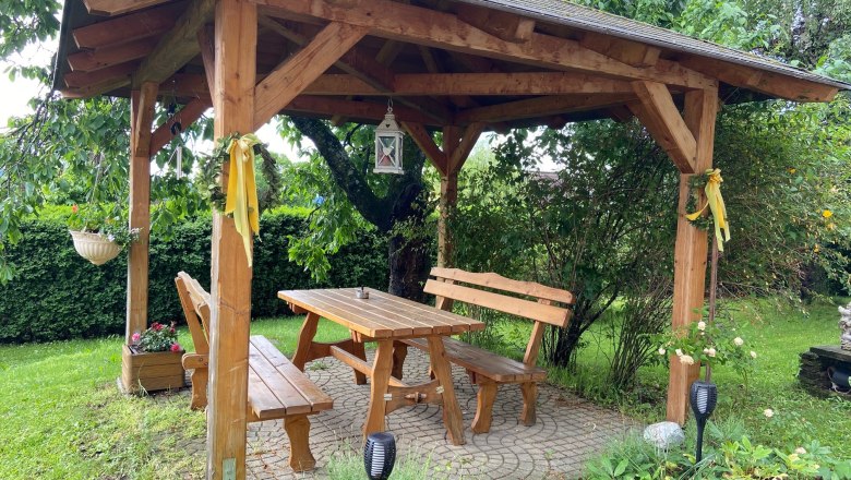 Holzlaube im Garten mit Tisch und Bänken, umgeben von Pflanzen.