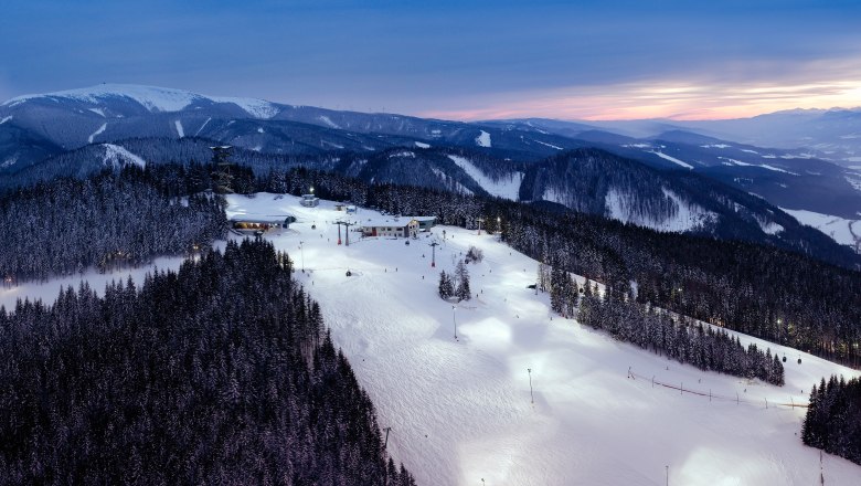 Beleuchtete Skipiste am Semmering bei Dämmerung mit schneebedeckten Bäumen und Bergen im Hintergrund.
