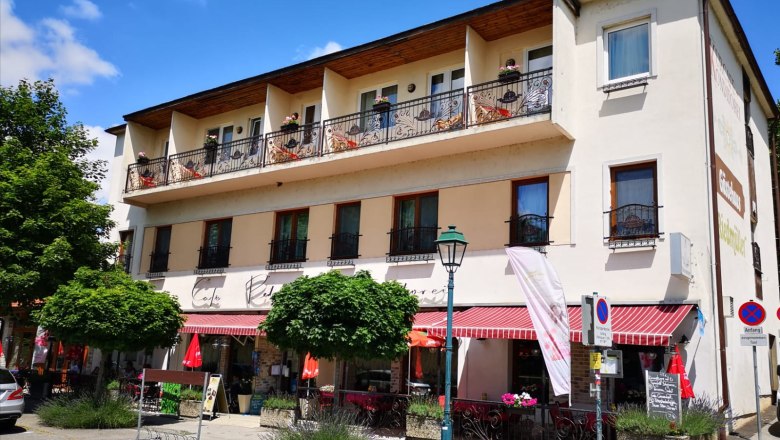 Aussenaufnahme, © Hotel- Kur-Kaffee-Konditorei & Restaurant "König Stefan" Außenansicht eines Cafés mit Balkon und Markise, umgeben von Bäumen und Straßenschildern.