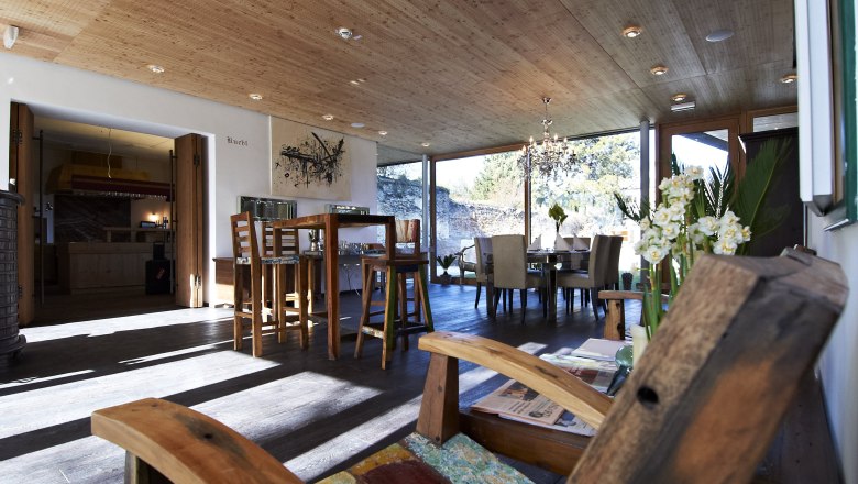 Ein moderner Fr&uuml;hst&uuml;cksraum mit Holzm&ouml;beln, gro&szlig;en Fenstern und einem Kronleuchter. Im Vordergrund ein Holzstuhl, im Hintergrund ein Esstisch.