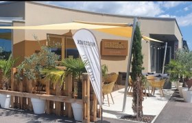 Außenansicht eines Cafés mit Terrasse, Sonnensegeln und Pflanzen.