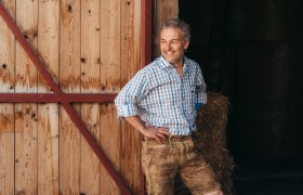 Mann in kariertem Hemd und Lederhose lehnt an Holzwand, l&auml;chelt.