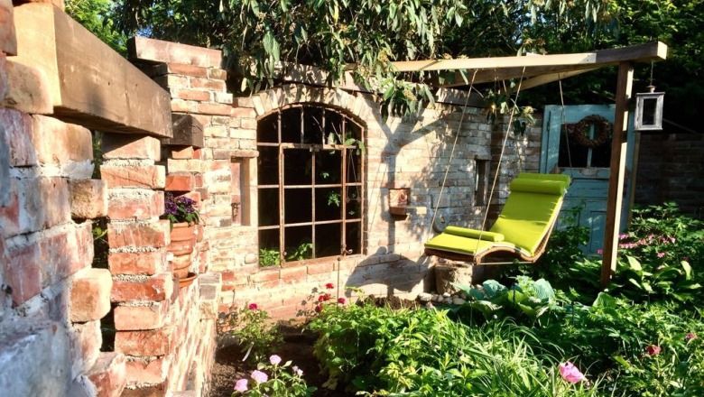Ein gemütlicher Garten mit einer alten Backsteinmauer, einem Fensterbogen und einer grünen Liege unter einem Holzrahmen.