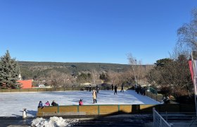 Eislaufplatz mit Menschen und Pinguinfigur, umgeben von Bäumen und Hügeln unter blauem Himmel.