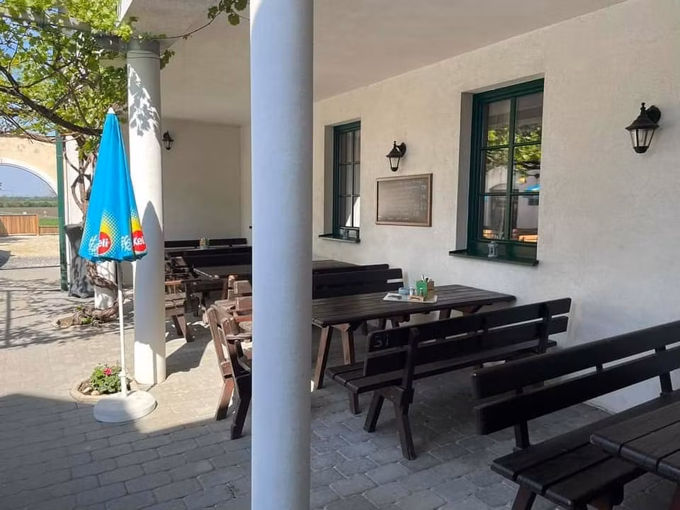 Ein leerer Gastgarten mit Holzbänken und Tischen, einem Sonnenschirm und einer weißen Wand mit Fenstern.