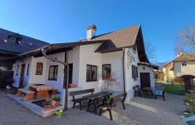 Ein traditionelles Haus mit Holzbänken und Tischen im Garten, umgeben von blühenden Pflanzen und Bäumen unter einem klaren blauen Himmel.