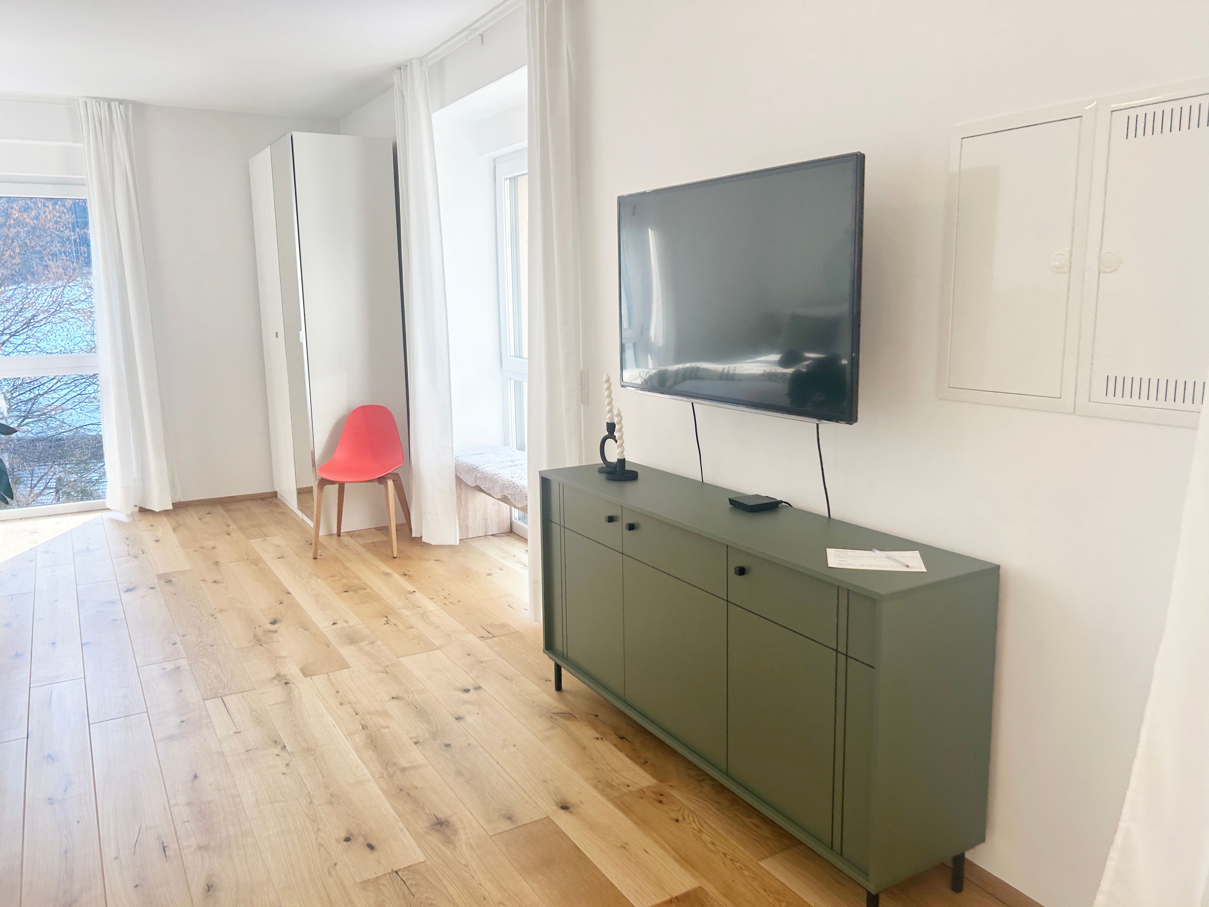 Modernes Wohnzimmer mit Holzboden, grünem Sideboard, Wandfernseher und rotem Stuhl vor einem Spiegel.