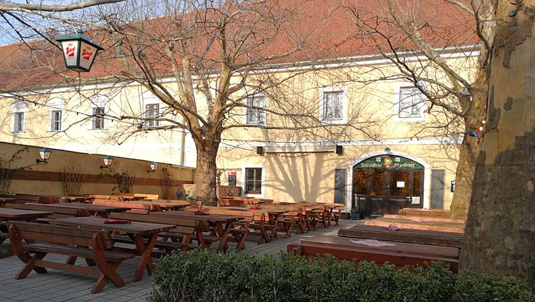 Biergarten mit Holzbänken und Tischen vor einem gelben Gebäude.