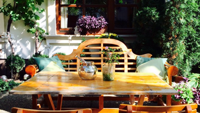 Gem&uuml;tlicher Gartenbereich mit Holztisch und St&uuml;hlen, umgeben von Pflanzen und Weinreben.