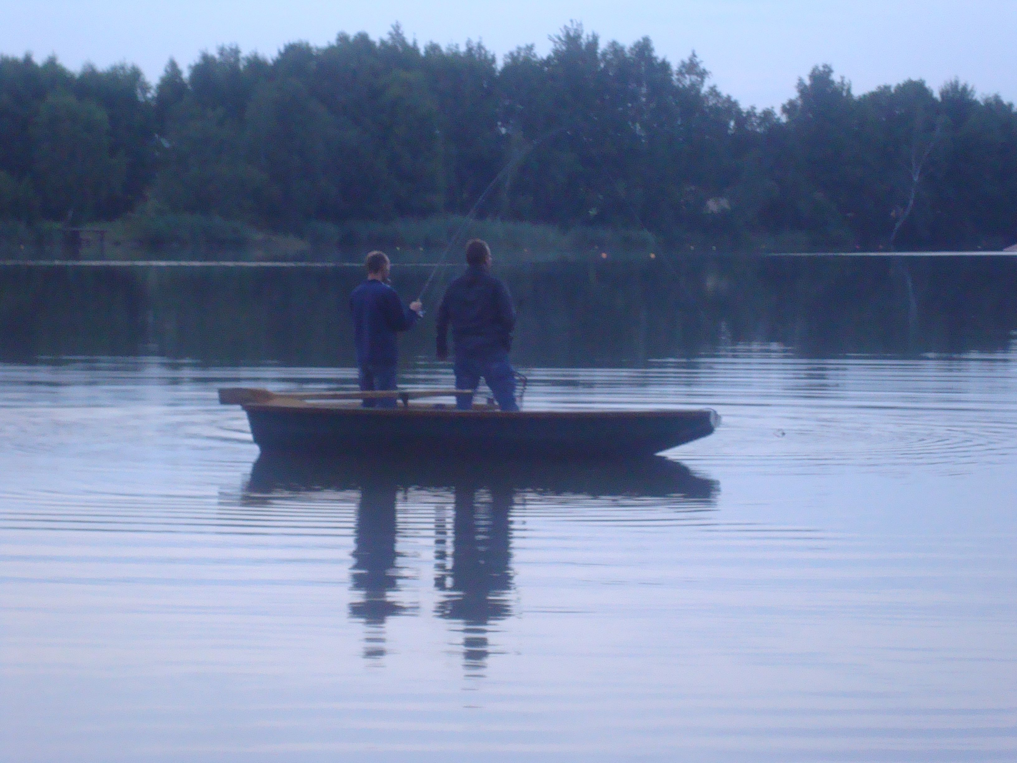 Zwei Personen angeln auf einem Boot auf einem ruhigen See, umgeben von Bäumen.