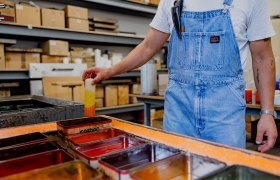 Person in Latzhose taucht Kerze in Farbbad.