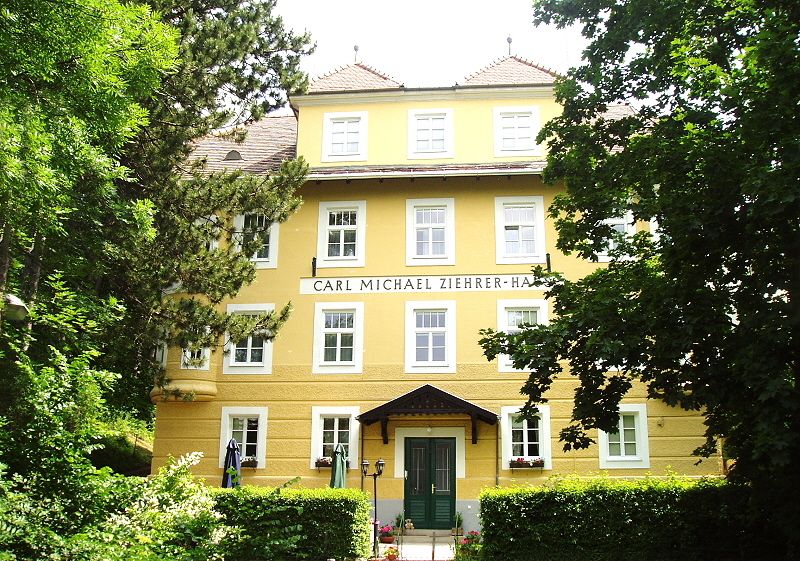 Frontansicht des Carl Michael Ziehrer-Hauses, ein gelbes Gebäude mit mehreren Fenstern und einem grünen Eingang, umgeben von Bäumen.