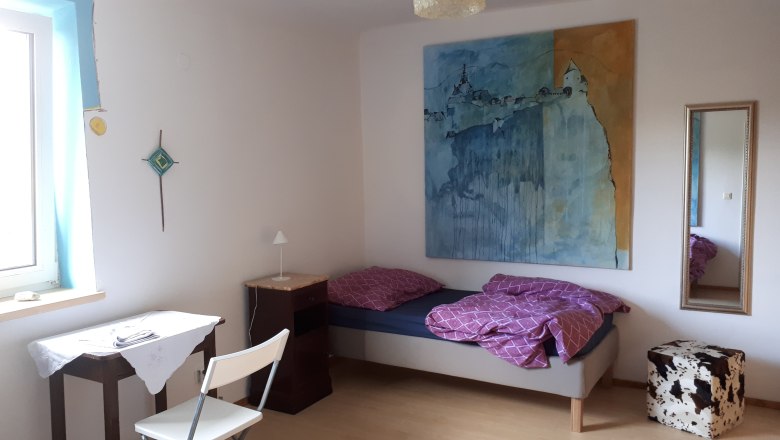 Ein einfach eingerichtetes Zimmer mit Bett, Tisch, Stuhl und großem Gemälde an der Wand.