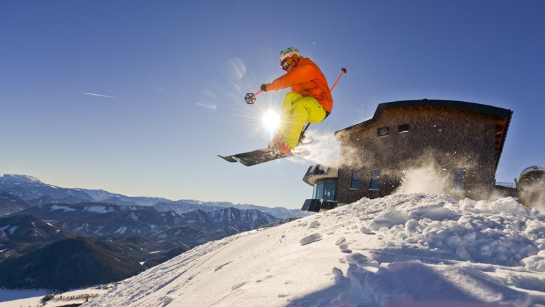 Ein Skifahrer springt vor dem Terzerhaus auf der Gemeindealpe in die Luft, umgeben von schneebedeckten Bergen und blauem Himmel.