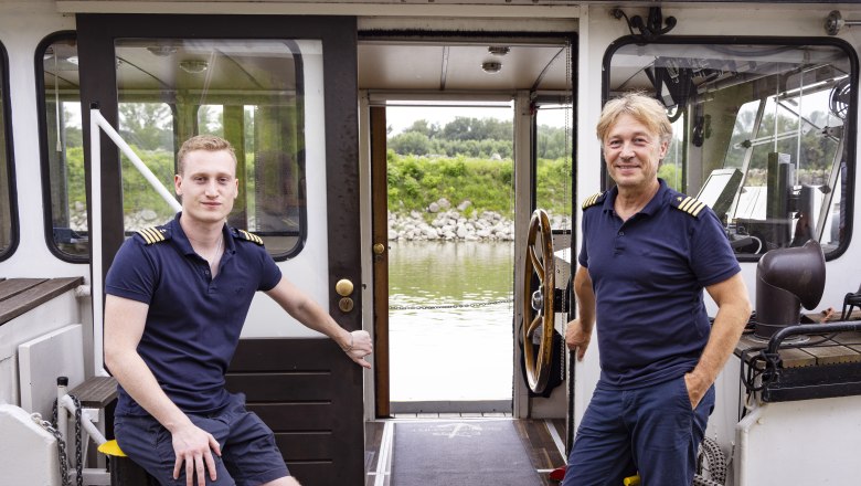 Zwei M&auml;nner in Marineuniform auf einem Schiff, mit Blick auf einen Fluss und gr&uuml;ne Uferlandschaft.