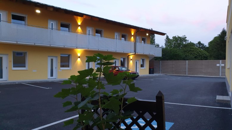 Au&szlig;enansicht eines zweist&ouml;ckigen Geb&auml;udes mit gelber Fassade und beleuchtetem Balkon, davor ein geparkter roter Wagen und Pflanzen im Vordergrund.