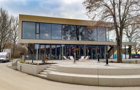 Moderne zweist&ouml;ckige Geb&auml;ude mit Glasfassade und Terrasse, umgeben von B&auml;umen.