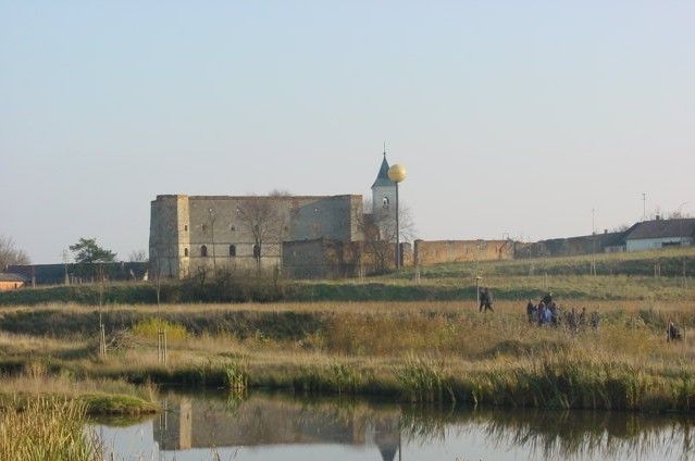 Ruine mit Turm und goldener Kugel in einer ländlichen Landschaft mit Teich im Vordergrund.