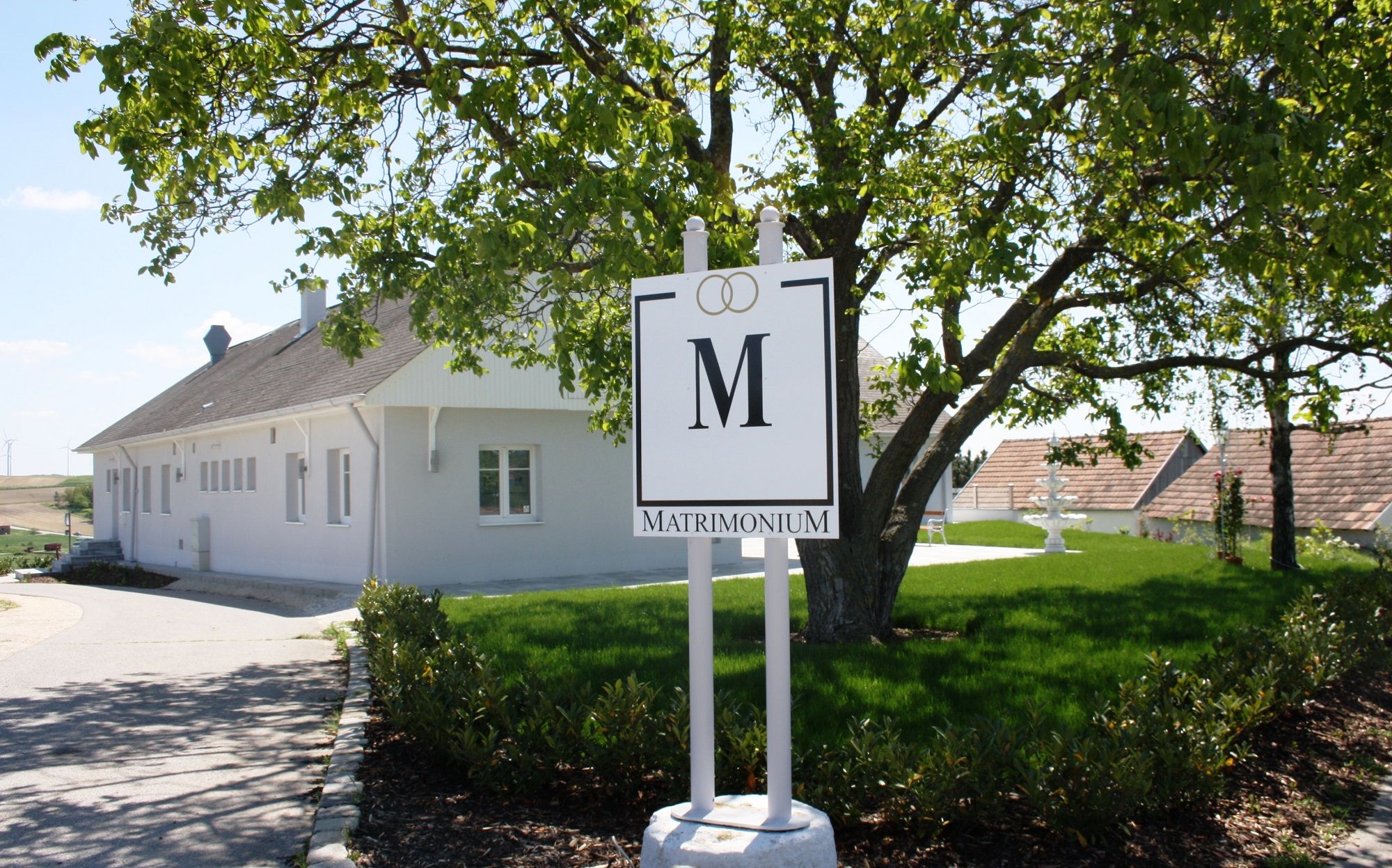 Ein weißes Gebäude mit einem Schild "Matrimonium" im Vordergrund, umgeben von grüner Landschaft.