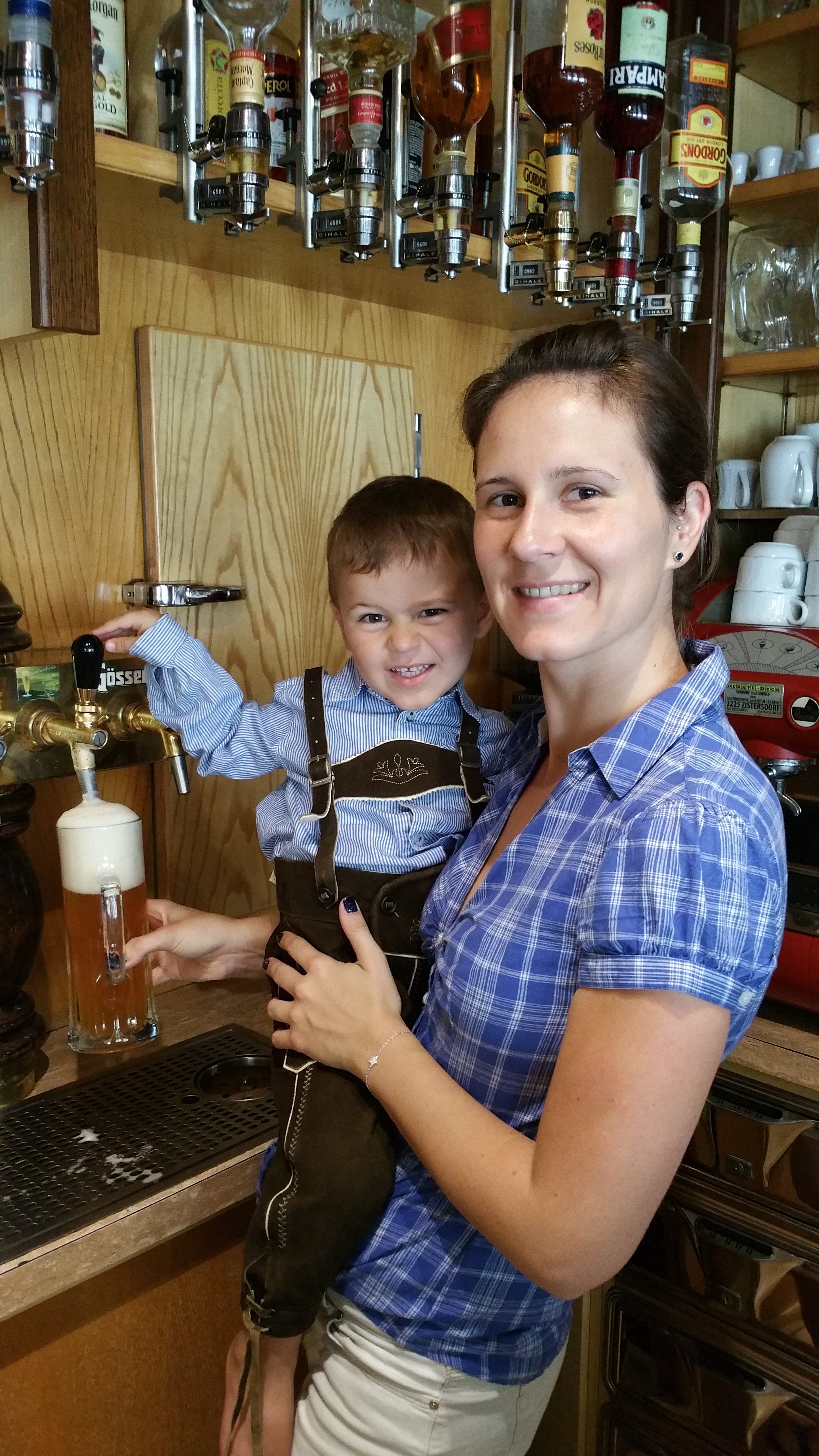Eine Frau hält ein Kind in Lederhose an einem Bierzapfhahn. Im Hintergrund sind Spirituosenflaschen zu sehen.