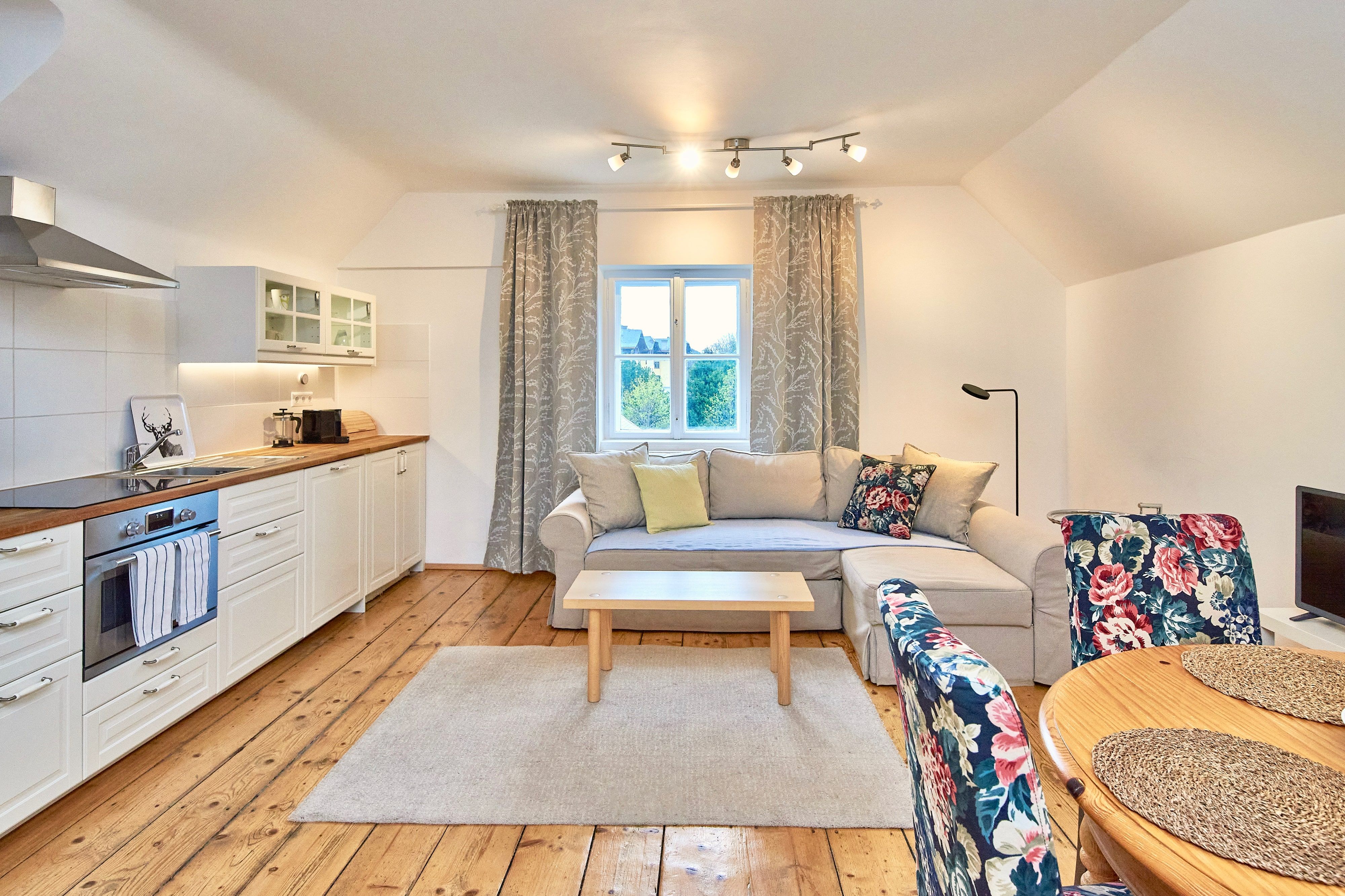 Gemütliches Wohnzimmer mit offener Küche, Holzboden, beiger Couch, floral gemusterten Stühlen und einem Fenster mit Vorhängen.