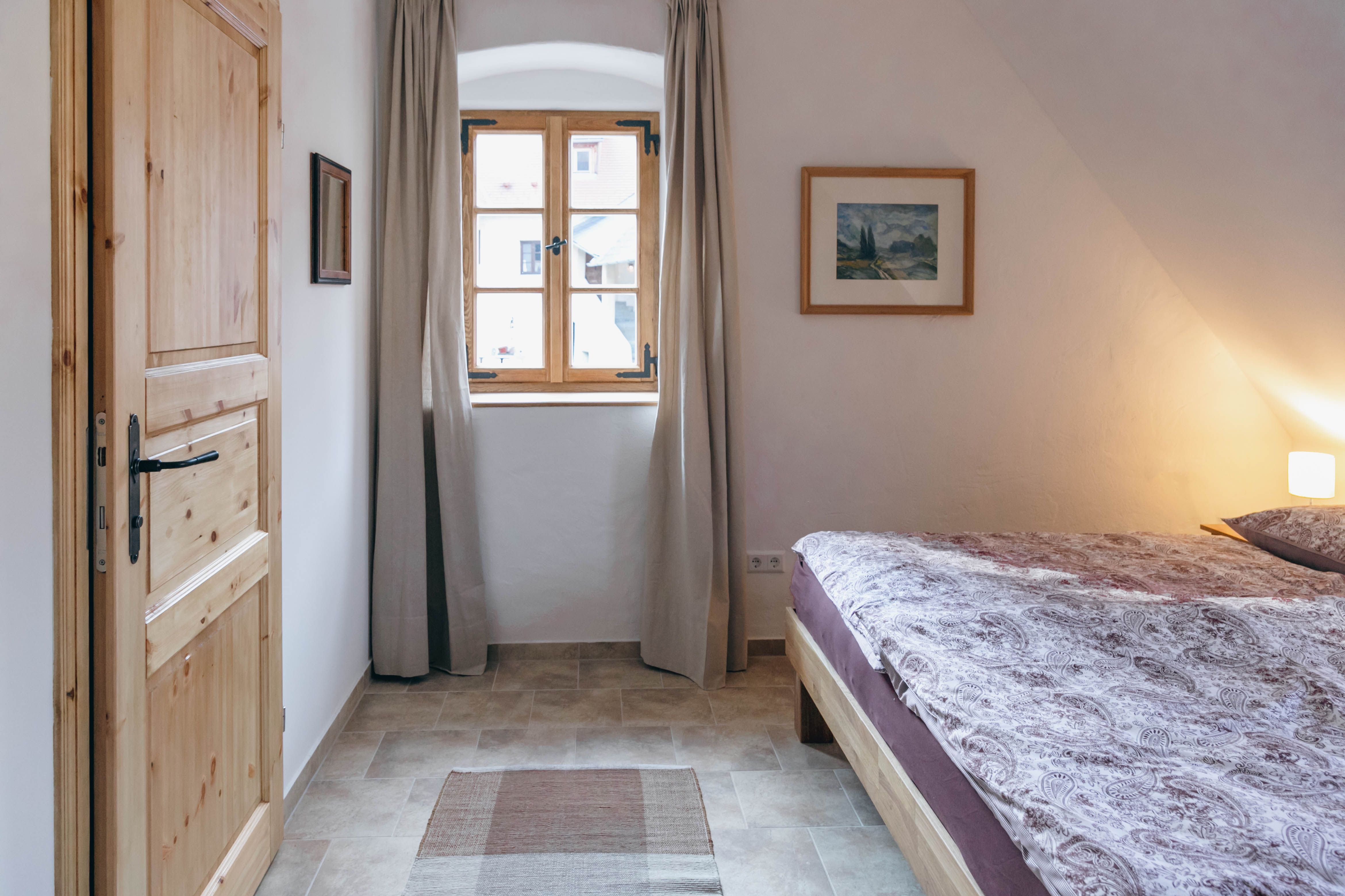 Ein gemütliches Schlafzimmer mit Holztür, Fenster mit Vorhängen, Doppelbett und Wandbild.