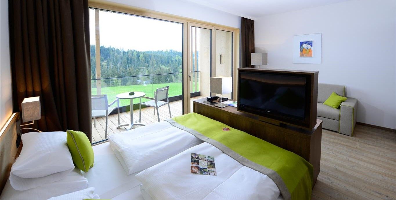 Modernes Hotelzimmer mit Doppelbett, Fernseher, Sofa und Balkon mit Waldblick.
