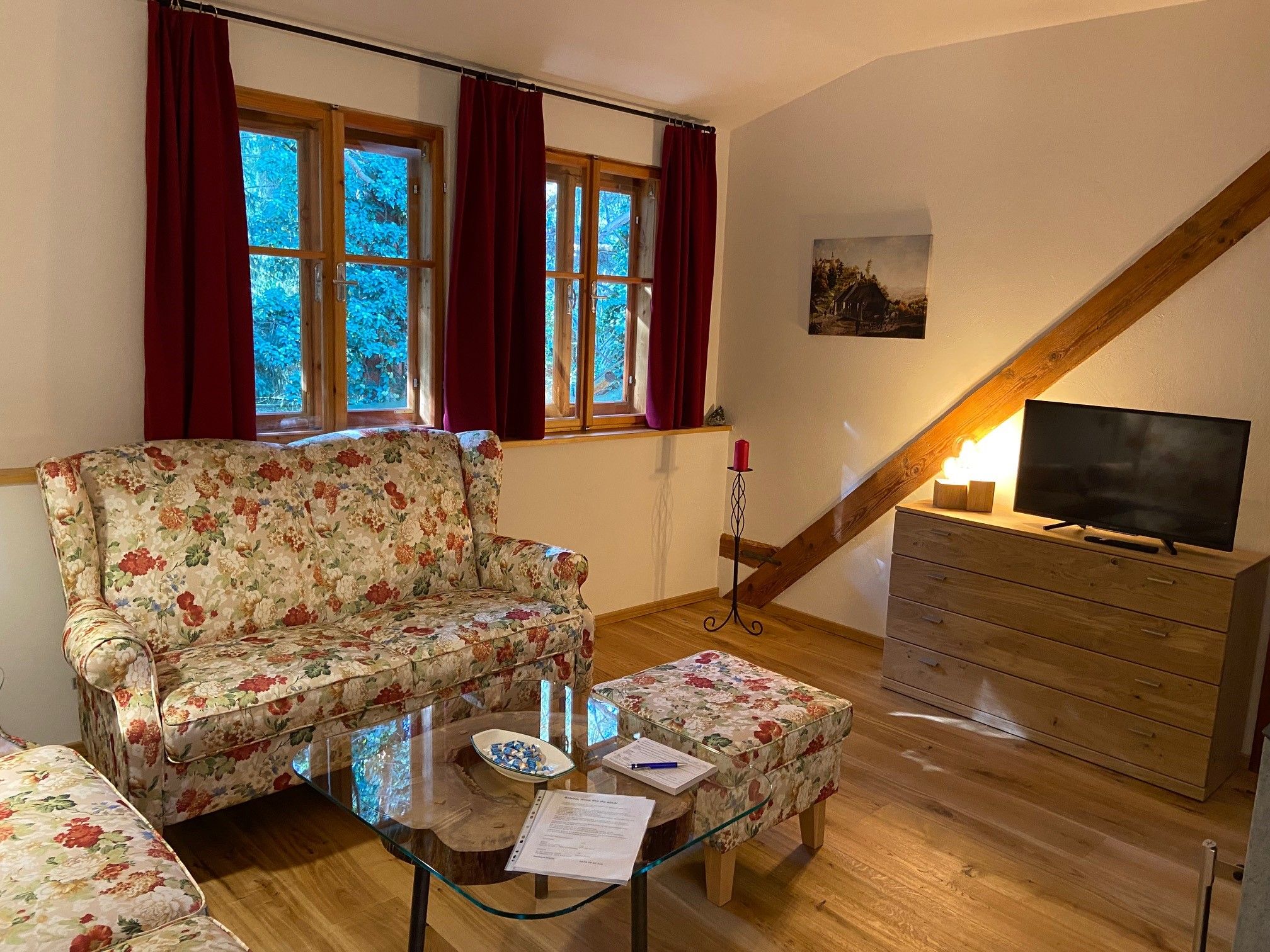 Gemütliches Wohnzimmer mit floralen Sofas, Holzmöbeln und einem Fernseher. Rote Vorhänge und ein Bild an der Wand.