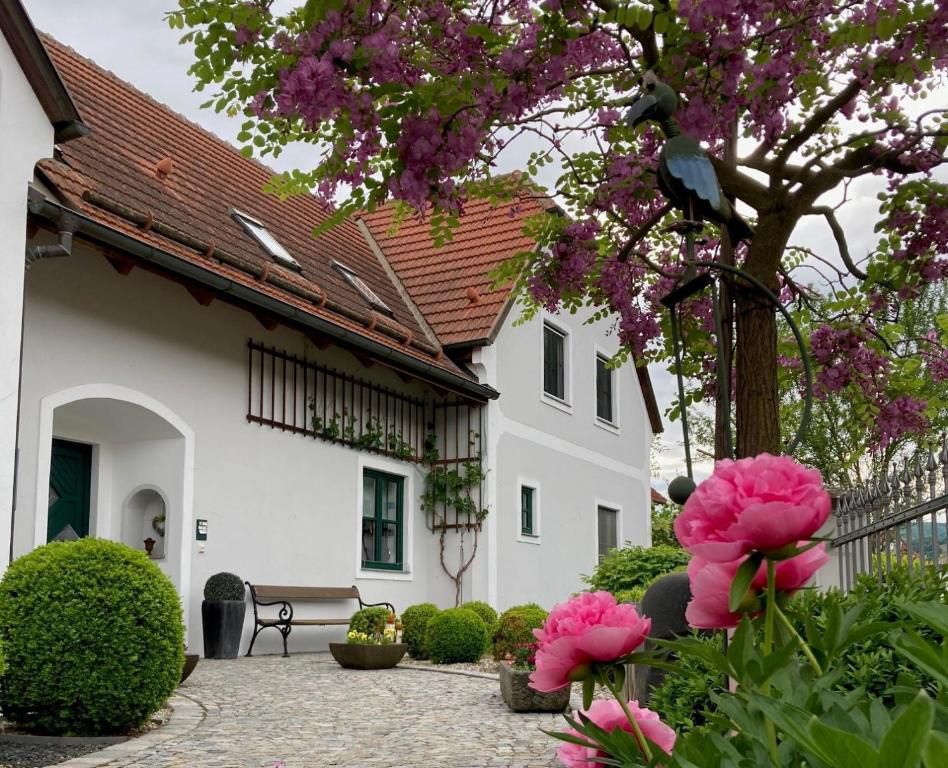 Ein Landhaus mit roten Dachziegeln, umgeben von blühenden Bäumen und rosa Blumen im Vordergrund.