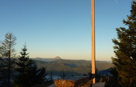 Gipfelkreuz am T&uuml;rnitzer H&ouml;ger mit Holzb&auml;nken und Bergpanorama im Hintergrund.