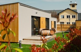 Moderne Appartements mit Holzverkleidung und Weinf&auml;ssern auf der Terrasse, umgeben von gr&uuml;nem Rasen und einem Zaun.