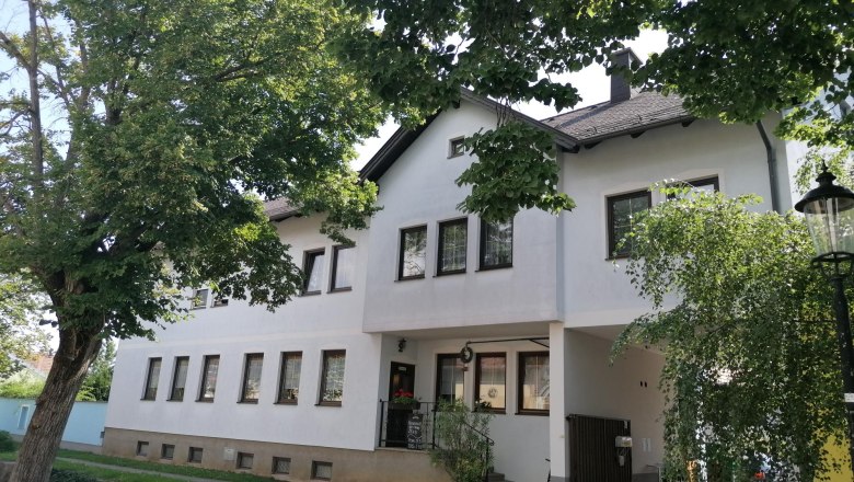 Ein wei&szlig;es Geb&auml;ude mit mehreren Fenstern, umgeben von B&auml;umen und einem Laternenpfahl.