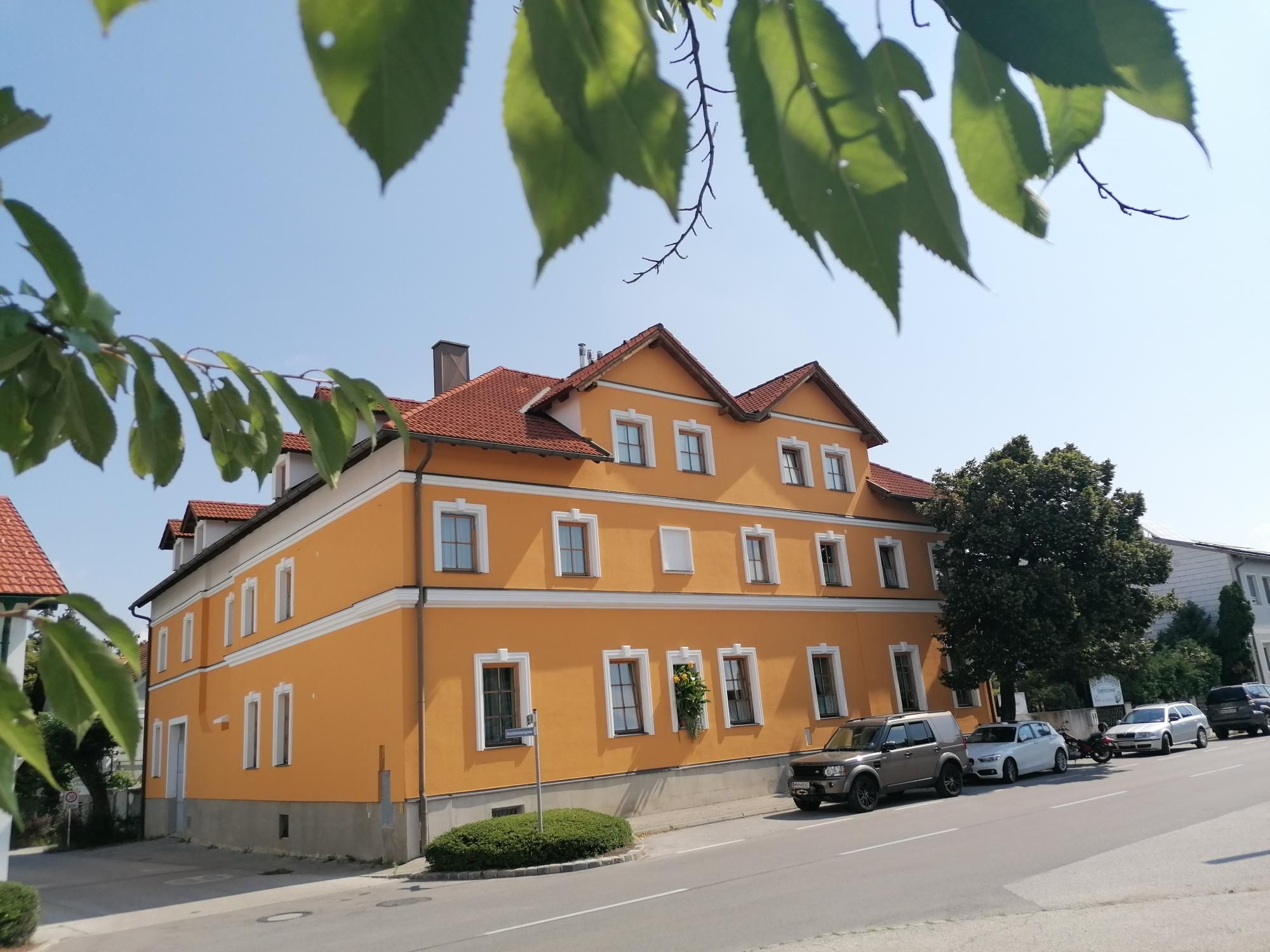 Gelbes Gebäude mit roten Dächern, umgeben von Bäumen und parkenden Autos.