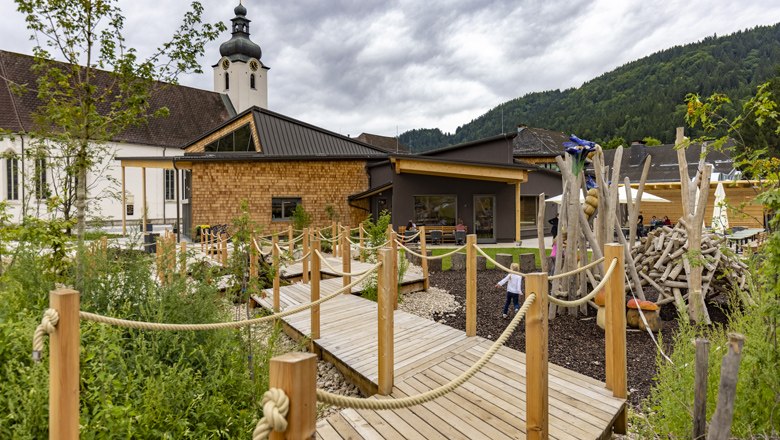Ein modernes Geb&auml;ude mit Holzverkleidung und Spielplatz, umgeben von gr&uuml;ner Landschaft und Bergen im Hintergrund.