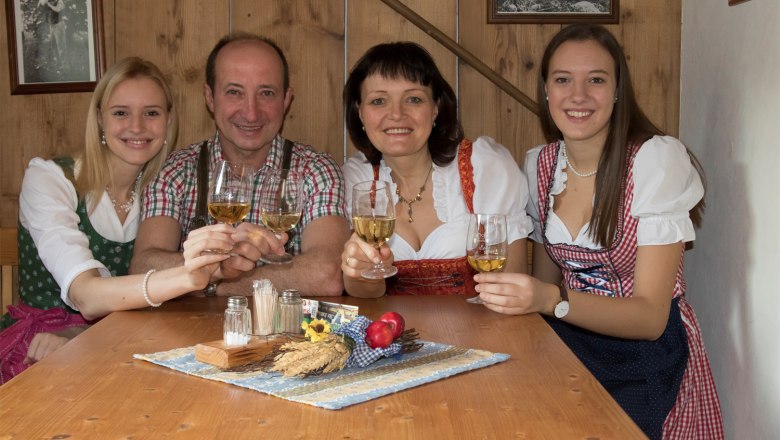 Eine Familie in traditioneller Kleidung st&ouml;&szlig;t mit Weingl&auml;sern an einem Holztisch an.
