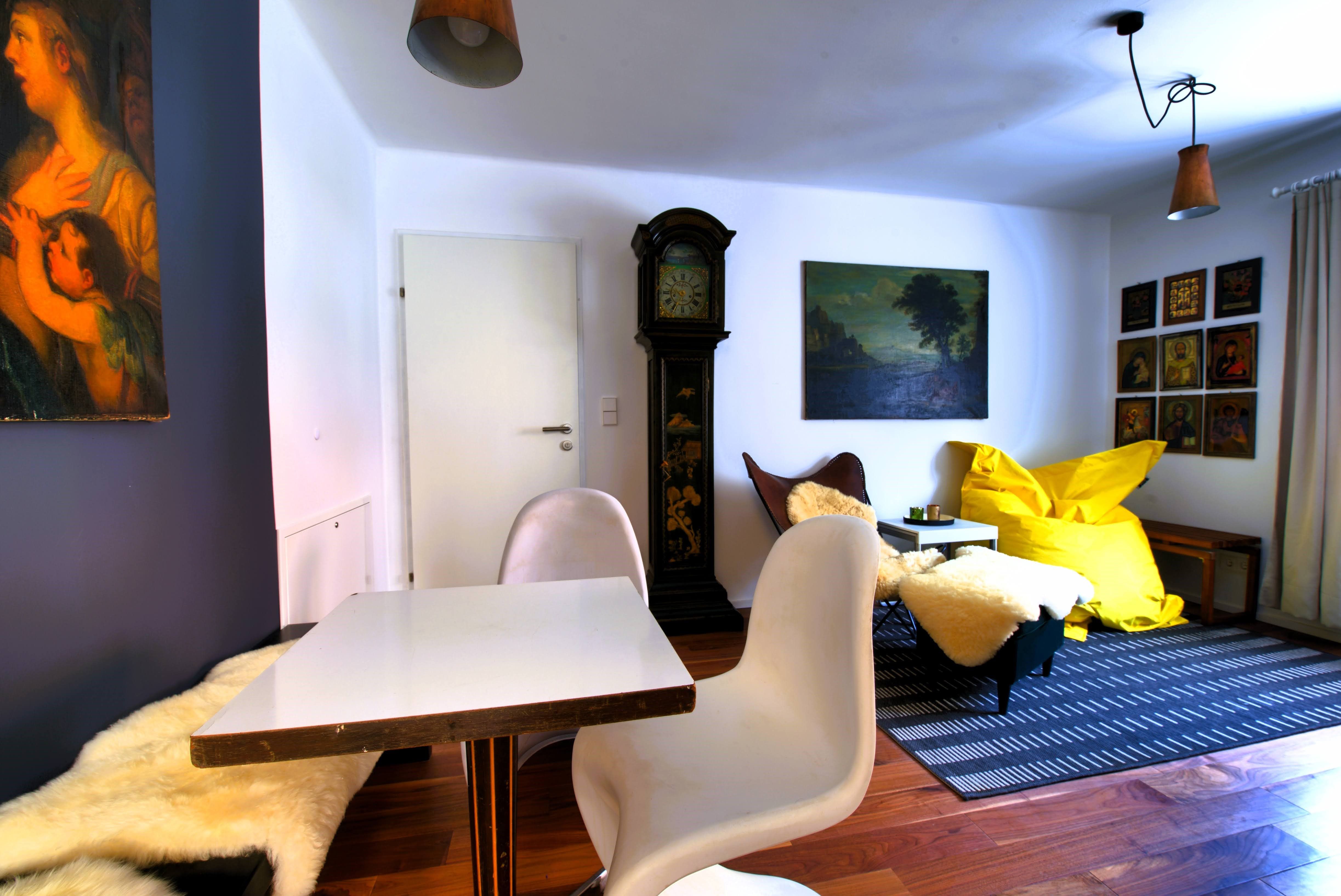 Wohnzimmer mit Tisch, Stühlen, Standuhr, Gemälden und gelbem Sitzsack.