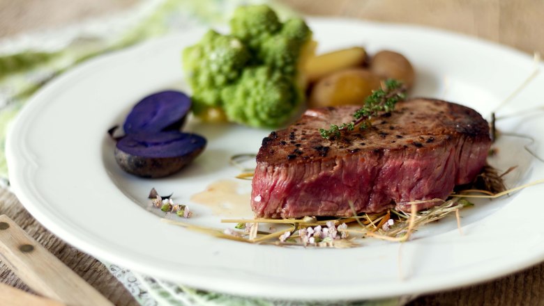Ein St&uuml;ck Steak auf einem wei&szlig;en runden Teller mit Gem&uuml;se, Kartoffeln und Kr&auml;utern.