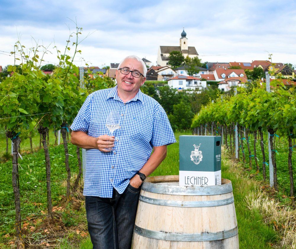 Mann im karierten Hemd mit Weinglas in einem Weinberg, im Hintergrund ein Dorf mit Kirche.